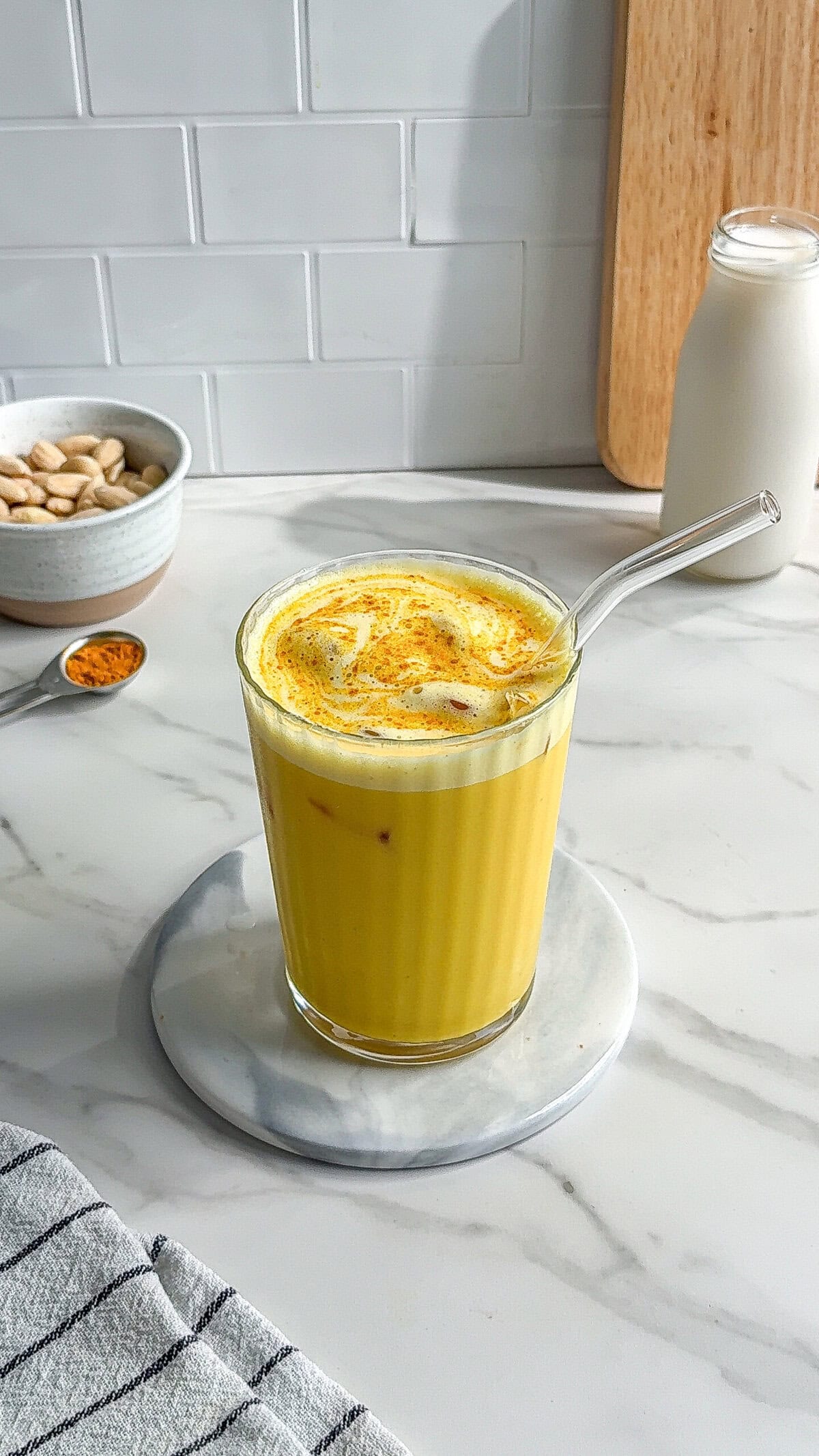 A glass of golden yellow vegan iced turmeric latte with a glass straw sits on a round marble coaster. In the background are a bowl of almonds, a spoon with turmeric powder, and a bottle of milk on a marble countertop.