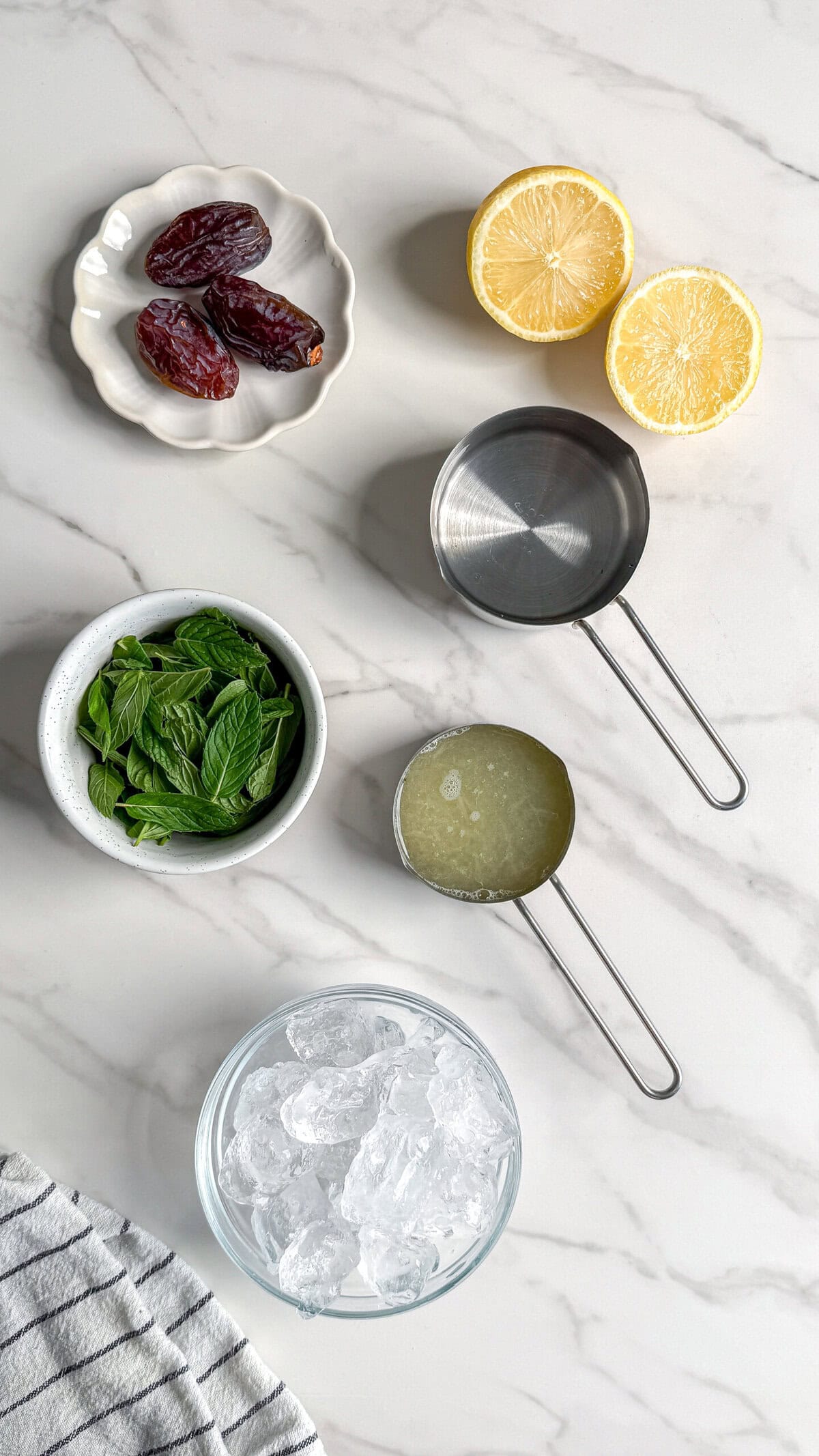 Top-down view of Paleo Frozen Mint Lemonades ingredients on marble: dates on a small plate, halved lemon, fresh mint leaves in a bowl, measuring cups of lemon juice and water, and a bowl of ice.