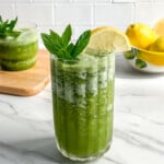 A glass of green smoothie, inspired by Paleo Frozen Mint Lemonades, is garnished with fresh mint leaves and a lemon slice on a white marble countertop. In the background are lemons in a bowl and another green smoothie on a wooden board.