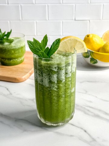 A glass of green smoothie, inspired by Paleo Frozen Mint Lemonades, is garnished with fresh mint leaves and a lemon slice on a white marble countertop. In the background are lemons in a bowl and another green smoothie on a wooden board.