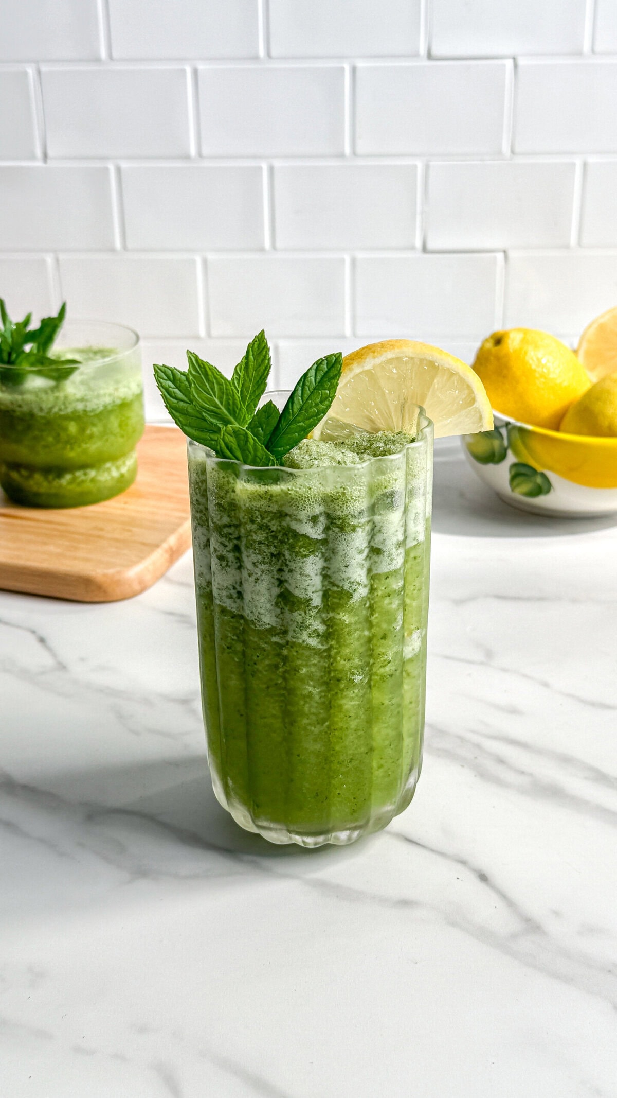 A glass of green smoothie, inspired by Paleo Frozen Mint Lemonades, is garnished with fresh mint leaves and a lemon slice on a white marble countertop. In the background are lemons in a bowl and another green smoothie on a wooden board.