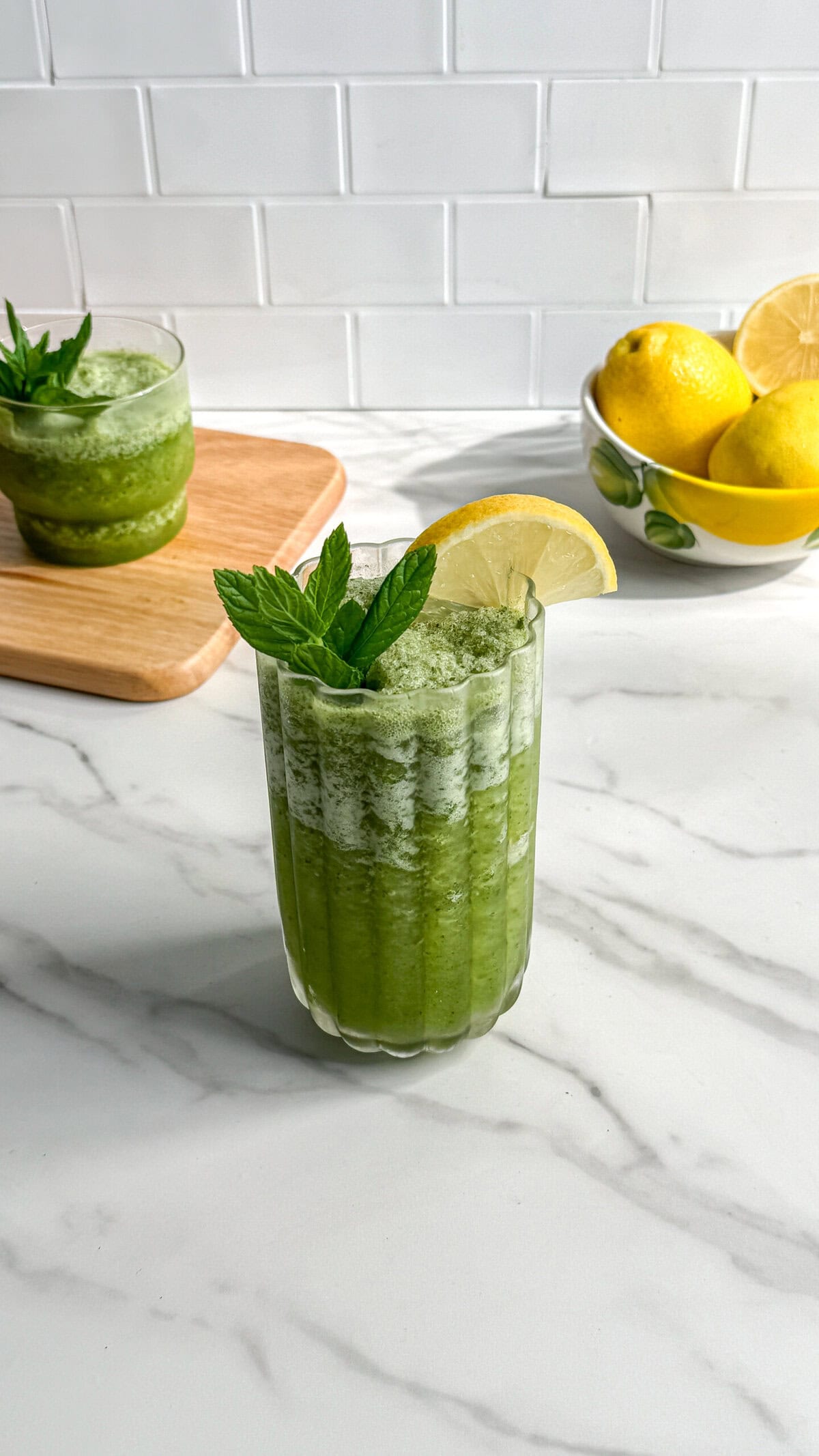 A glass of green smoothie, reminiscent of Paleo Frozen Mint Lemonades, is garnished with a lemon slice and mint sprig on a marble countertop. In the background, another glass, a wooden board, and a bowl of lemons sit against white tiles.