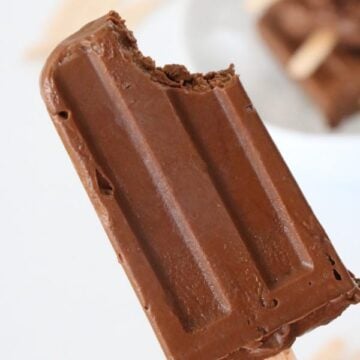 A hand holding a homemade fudgesicle with a bite taken out with a white background.