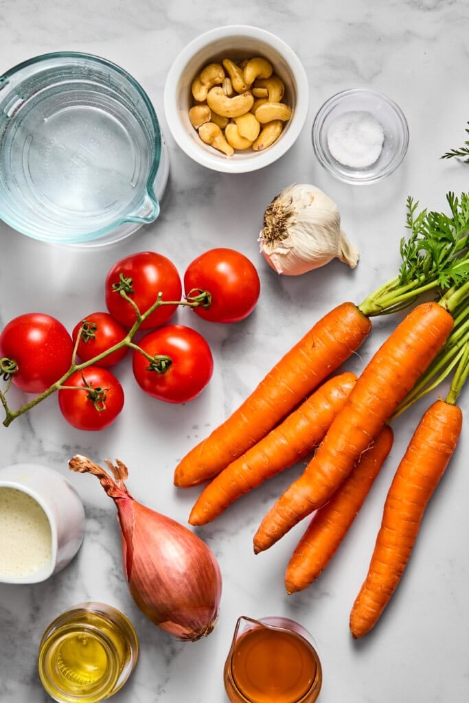 Carrots, vine tomatoes, garlic, shallot, cashews, a small bowl of salt, water, oil, cream, and honey are arranged on a marble countertop—the perfect ingredients for preparing a vibrant vegan carrot soup.