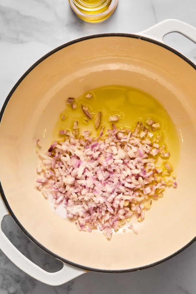 Chopped onions cooking in olive oil with salt in a large cream-colored pot on a marble countertop, forming the flavorful base for a vegan carrot soup. A small glass bowl of olive oil is visible at the top of the image.