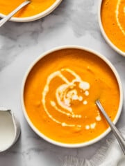 Three bowls of creamy vegan carrot soup with swirls of cream on top, each with a spoon. The bowls are on a white marble surface next to a small white pitcher and a textured white napkin.