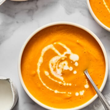 Three bowls of creamy vegan carrot soup with swirls of cream on top, each with a spoon. The bowls are on a white marble surface next to a small white pitcher and a textured white napkin.