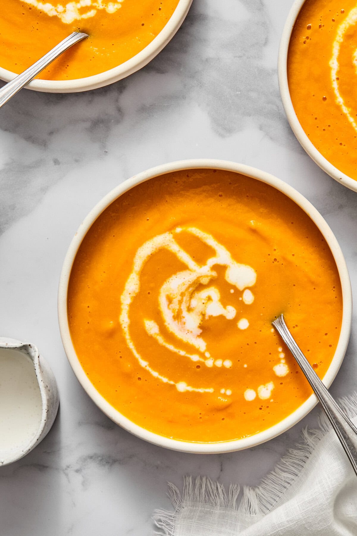 Three bowls of creamy vegan carrot soup with swirls of cream on top, each with a spoon. The bowls are on a white marble surface next to a small white pitcher and a textured white napkin.