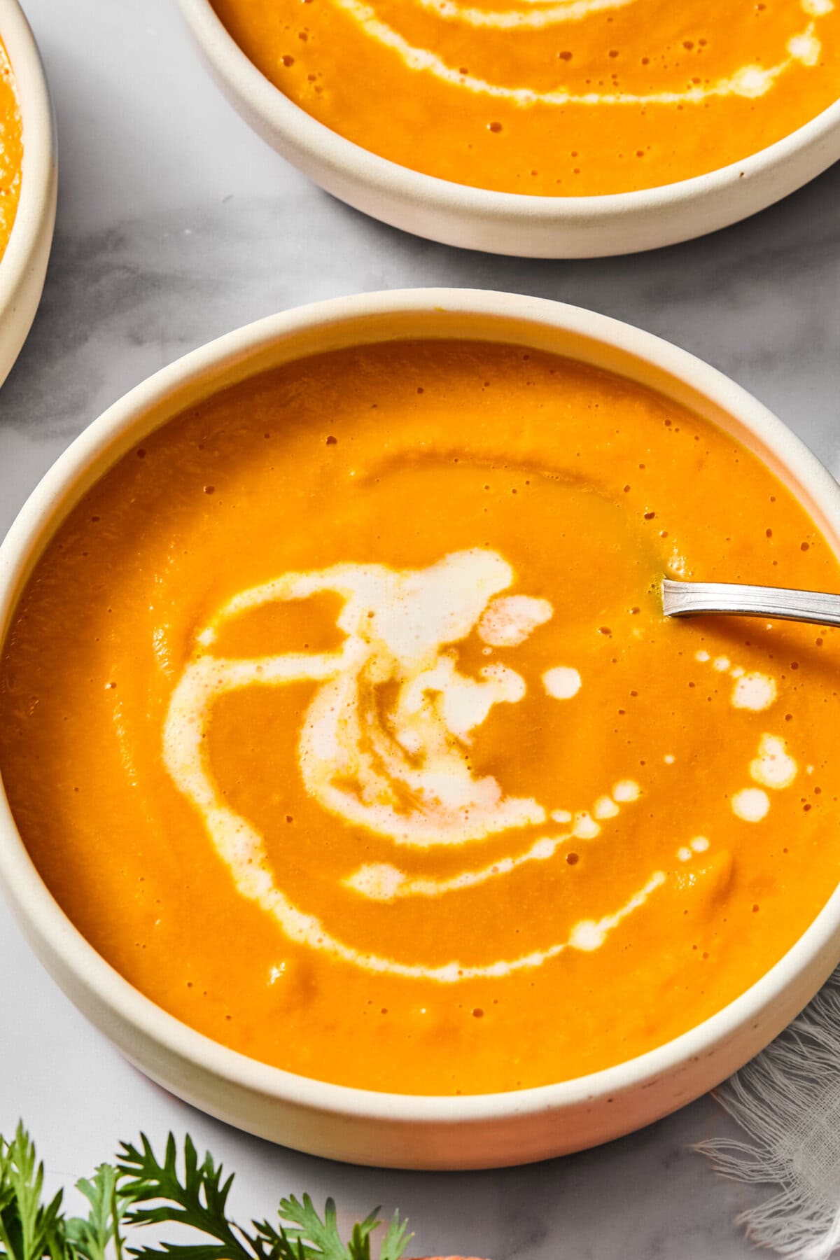 A bowl of creamy orange-colored vegan carrot soup with a swirl of cream on top. A spoon rests in the bowl, and fresh herbs are visible at the bottom of the image.