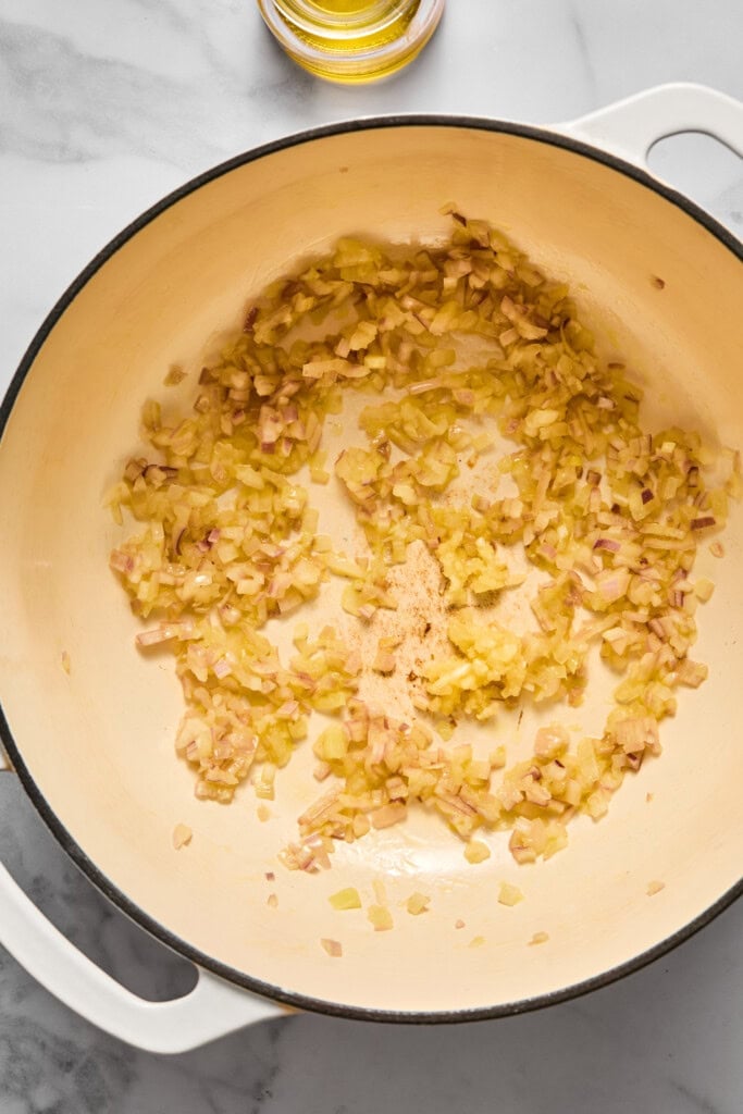 Chopped onions sautéing in a white pot on a marble countertop, with a small glass container of oil nearby—an inviting start to preparing a hearty vegan carrot soup.