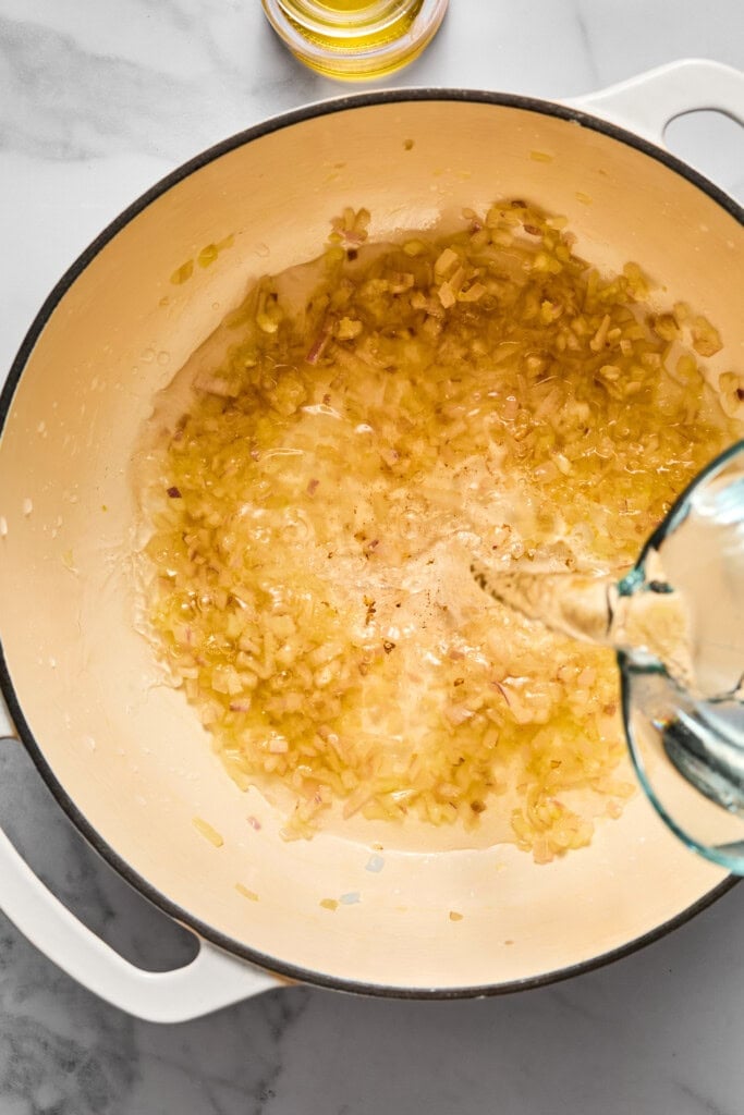 Chopped onions and garlic are sautéing in oil in a white pot, creating a flavorful base for vegan carrot soup, while liquid is being poured in, likely for cooking. A small bowl of oil sits nearby on the marble countertop.