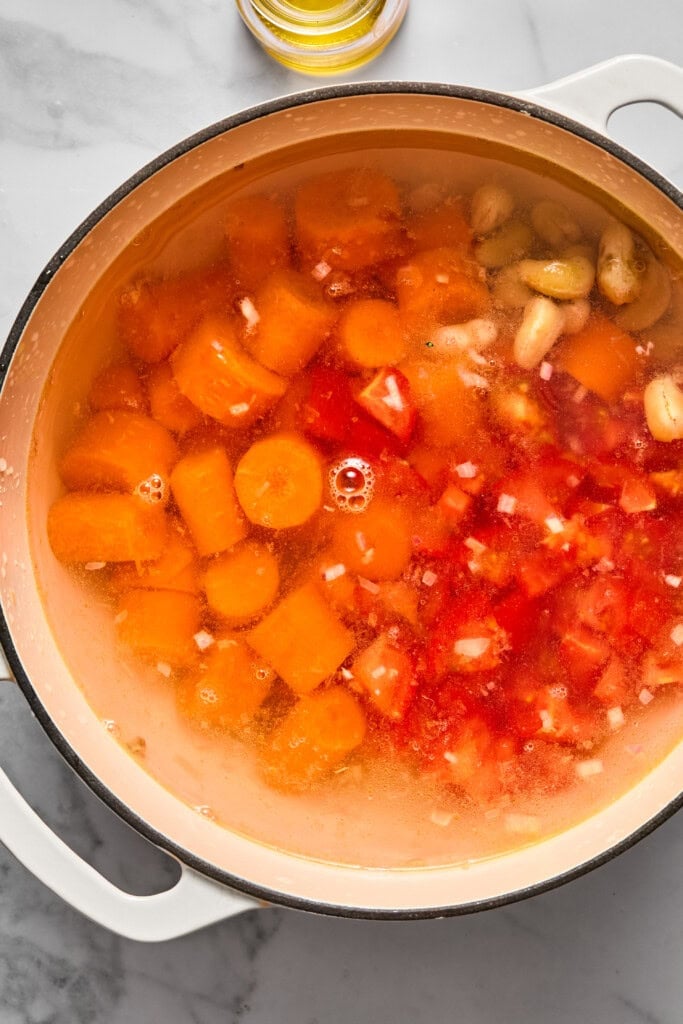 Chopped carrots, tomatoes, onions, and garlic cloves are simmering in a large white pot filled with water on a marble countertop, coming together to create a flavorful vegan carrot soup.