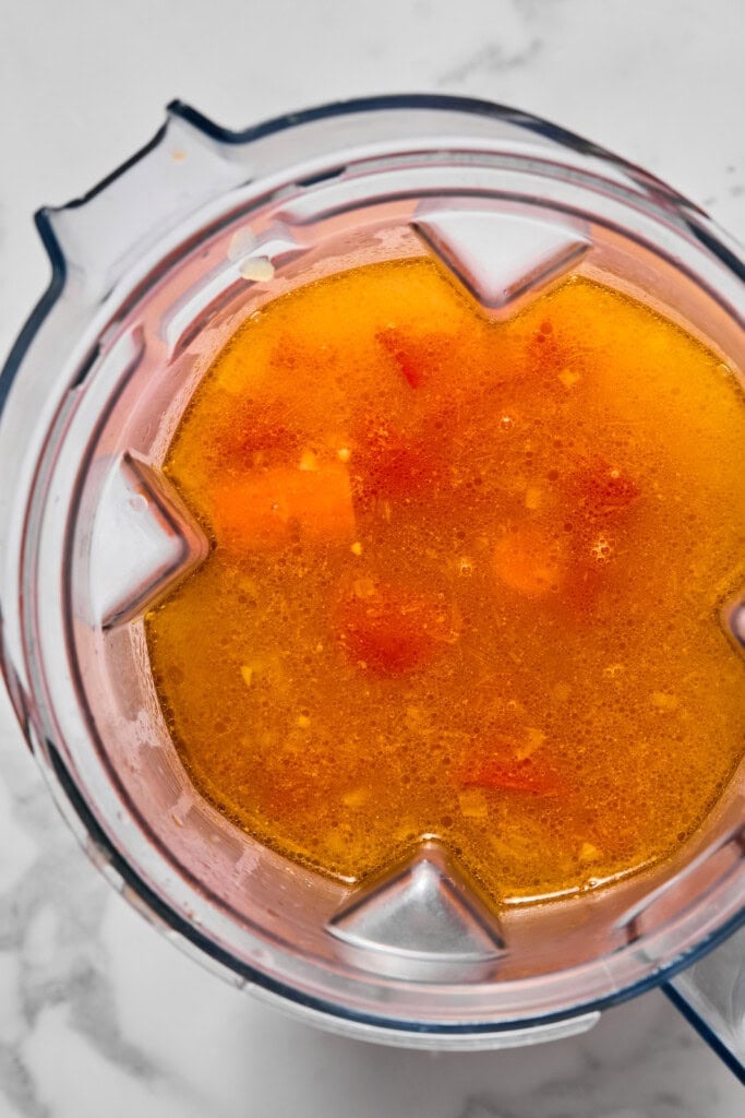 A blender filled with a chunky orange and red liquid mixture, including visible pieces of vegetables for a hearty vegan carrot soup, sits on a white countertop ready to be blended.