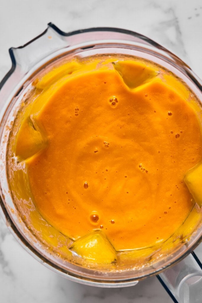Bright orange mango smoothie blending in a glass blender on a marble countertop, with visible chunks of mango around the edges—reminiscent of the vibrant color found in vegan carrot soup.