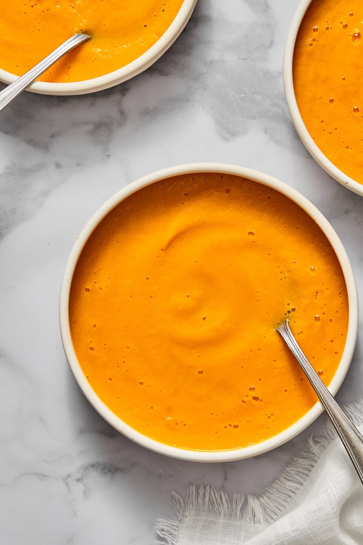 Three white bowls filled with creamy vegan carrot soup sit on a marble surface, each with a metal spoon. A white napkin is partially visible in the lower right corner.