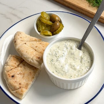 A plate with pita bread slices, a small bowl of creamy dip with herbs and a spoon, whole roasted carrots, and a side of green olives and pickles. Fresh dill is on a wooden board in the background.