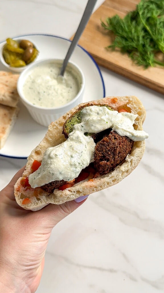 A hand holds a pita sandwich filled with falafel balls, diced tomatoes, and creamy white sauce. In the background, whole roasted carrots join pita, pickles, olives, a bowl of sauce, and fresh herbs on a wooden board.
