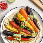 A plate with colorful whole roasted carrots on a swirl of hummus, topped with green sauce, chopped herbs, and pomegranate seeds. Next to the plate are a small bowl of pomegranate seeds, a jar of green sauce, and utensils.