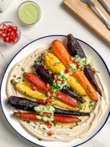 A plate with colorful whole roasted carrots on a swirl of hummus, topped with green sauce, chopped herbs, and pomegranate seeds. Next to the plate are a small bowl of pomegranate seeds, a jar of green sauce, and utensils.