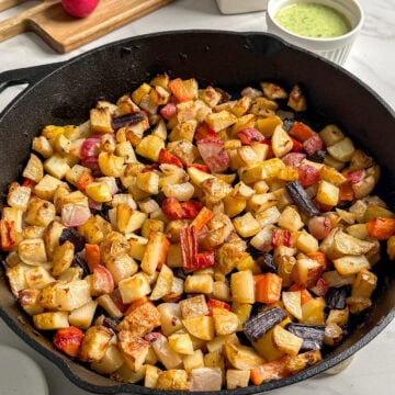 A cast iron skillet filled with roasted diced root vegetables sits on a white counter. Nearby are fresh radishes on a cutting board, a small pitcher of oil, and a bowl of green sauce. This vibrant root vegetable medley is perfect for any meal.