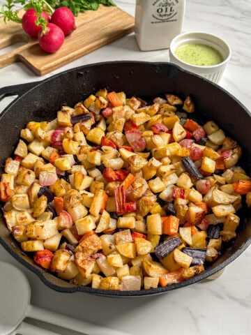 A cast iron skillet filled with roasted diced root vegetables sits on a white counter. Nearby are fresh radishes on a cutting board, a small pitcher of oil, and a bowl of green sauce. This vibrant root vegetable medley is perfect for any meal.