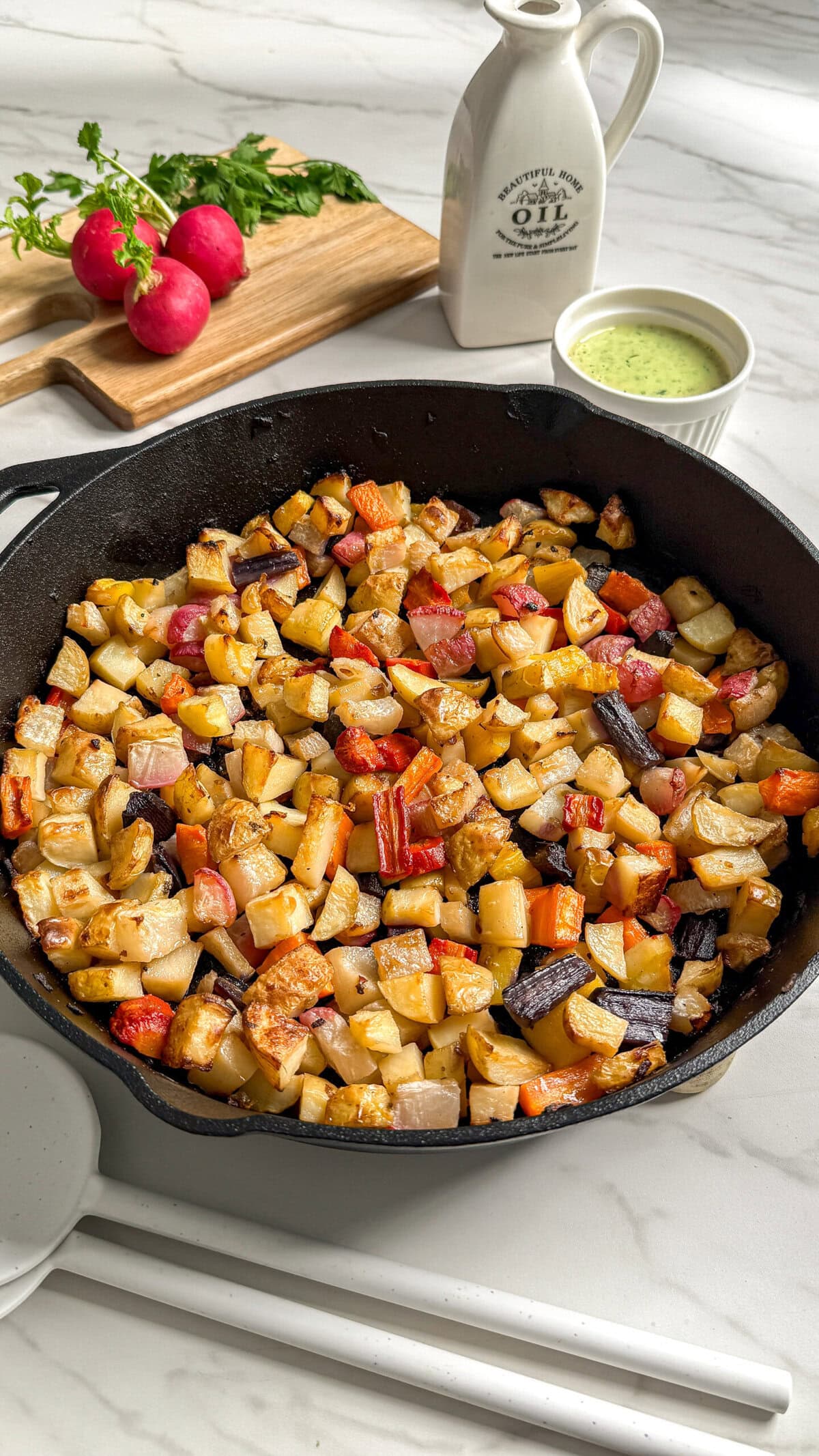 A cast iron skillet filled with roasted diced root vegetables sits on a white counter. Nearby are fresh radishes on a cutting board, a small pitcher of oil, and a bowl of green sauce. This vibrant root vegetable medley is perfect for any meal.