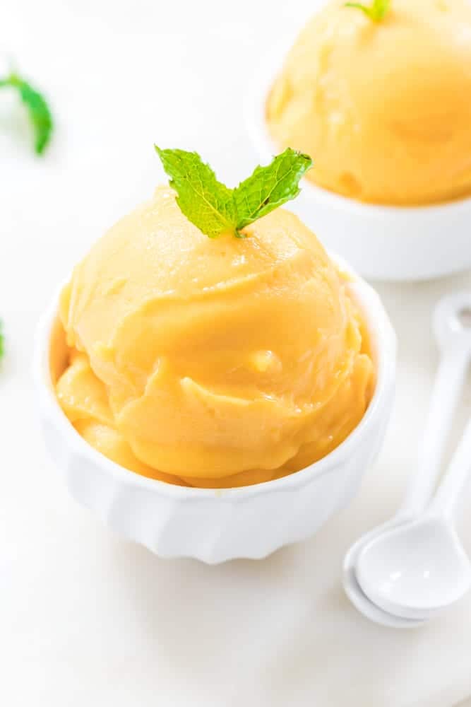 Homemade mango sorbet in a white bowl next to a white spoon with a white background.