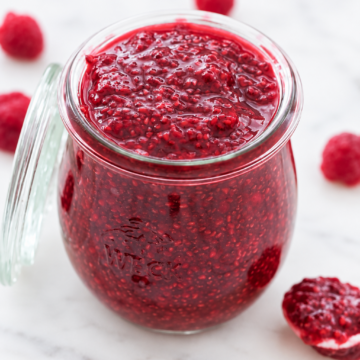 A glass jar filled with homemade raspberry chia jam sits on a white marble surface, with fresh raspberries scattered around.