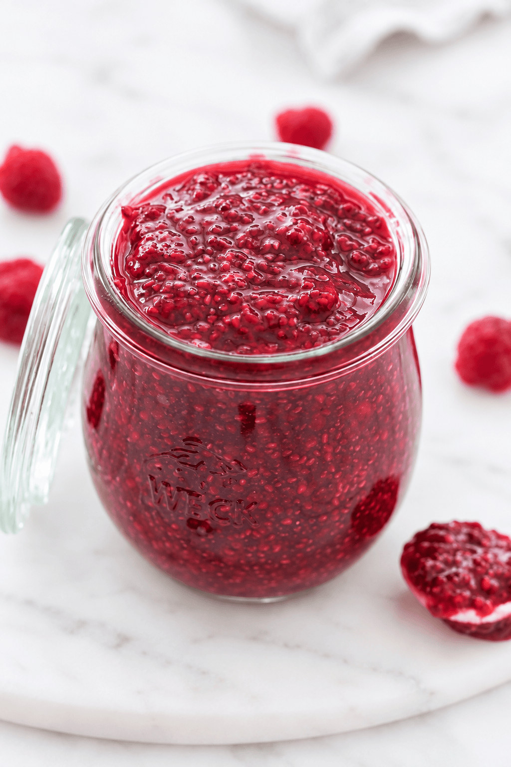 A glass jar filled with homemade raspberry chia jam sits on a white marble surface, with fresh raspberries scattered around.