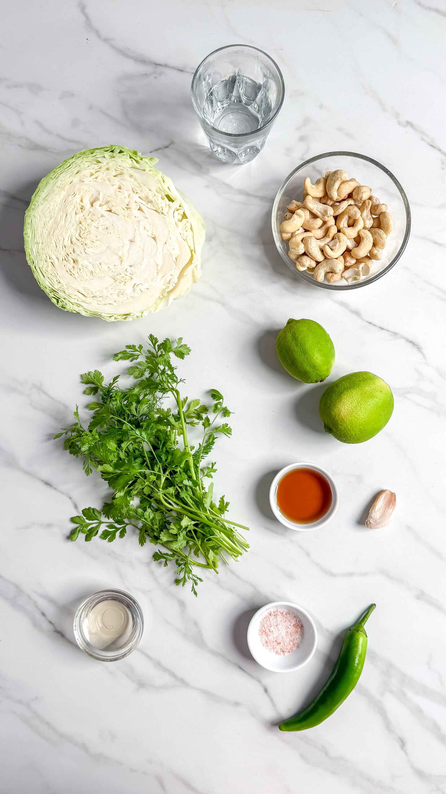 Ingredients for a fresh no mayo coleslaw on a marble surface, featuring half a cabbage, cashews, limes, cilantro, water, green chili, garlic, pink salt, tahini, and tamari sauce.