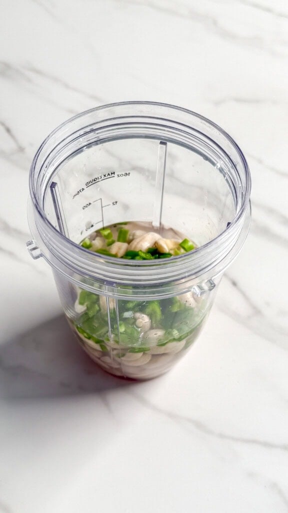 A clear blender cup on a white marble surface, filled with chopped green onions, mushrooms, and liquid—perfect for creating a flavorful base for a no mayo coleslaw or other fresh recipes.