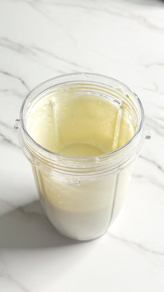 A clear blender cup containing a smooth, pale cream-colored no mayo coleslaw sauce or smoothie sits on a white marble surface.