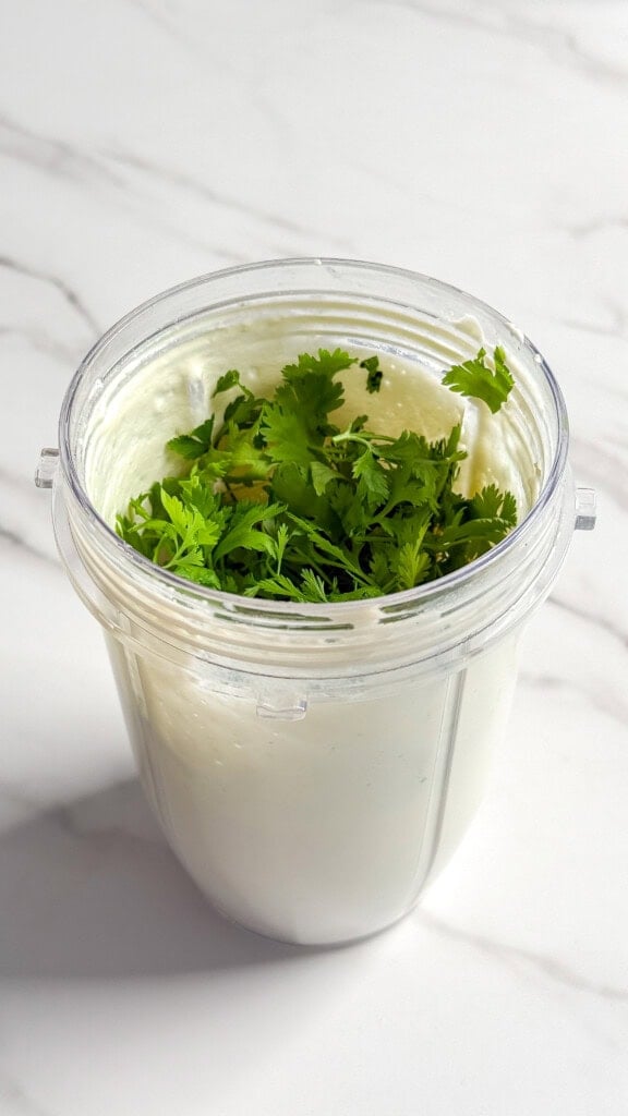 A blender cup filled with creamy white yogurt, perfect for making no mayo coleslaw, topped with fresh cilantro leaves and set on a white marble surface.