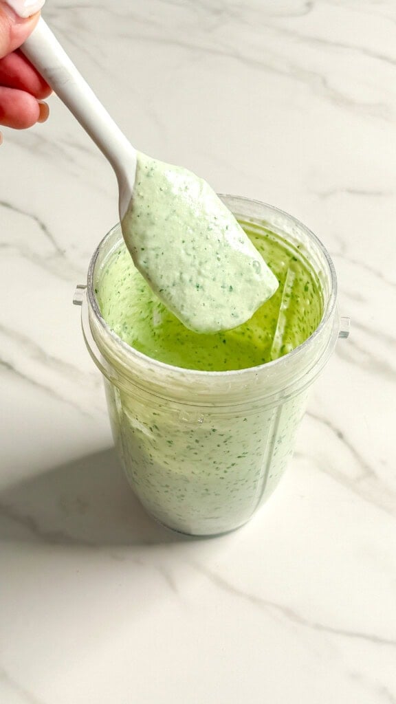 A spatula holds up a portion of creamy green no mayo coleslaw dressing above a clear blender cup, with more sauce inside, on a white marble countertop.