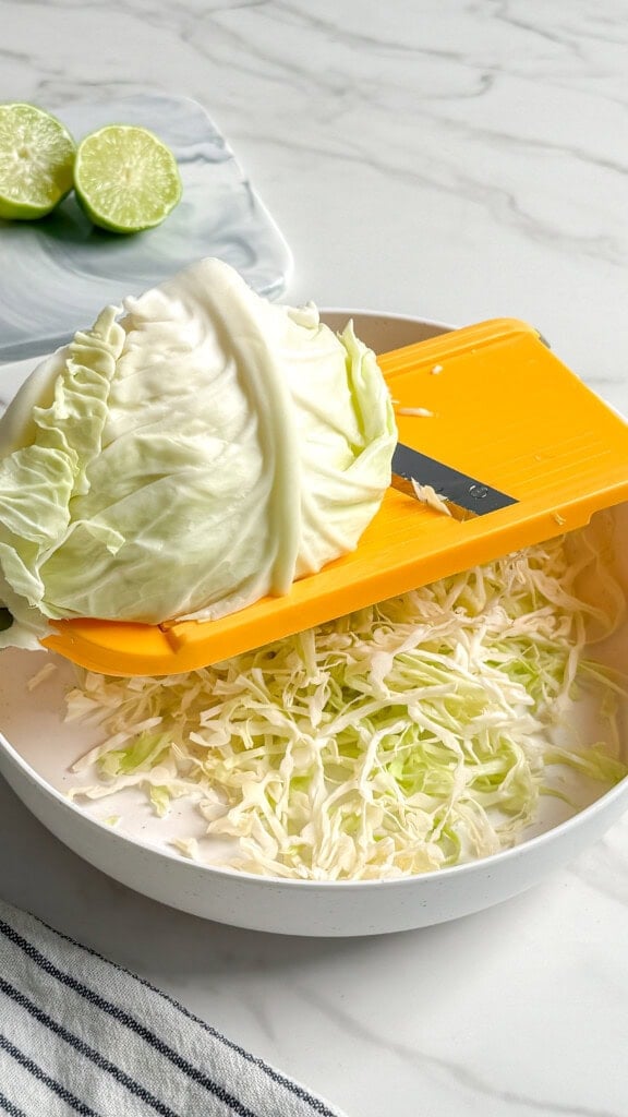 A head of cabbage is being sliced with a yellow mandoline slicer over a white bowl of shredded cabbage for a fresh, no mayo coleslaw. Two halved limes rest on a plate in the background atop a marble countertop.