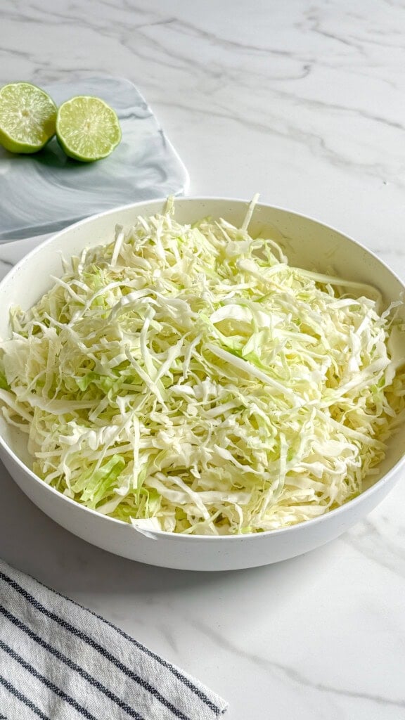 A white bowl filled with finely shredded cabbage for a no mayo coleslaw sits on a marble countertop, with a halved lime on a small plate and a striped cloth napkin nearby.