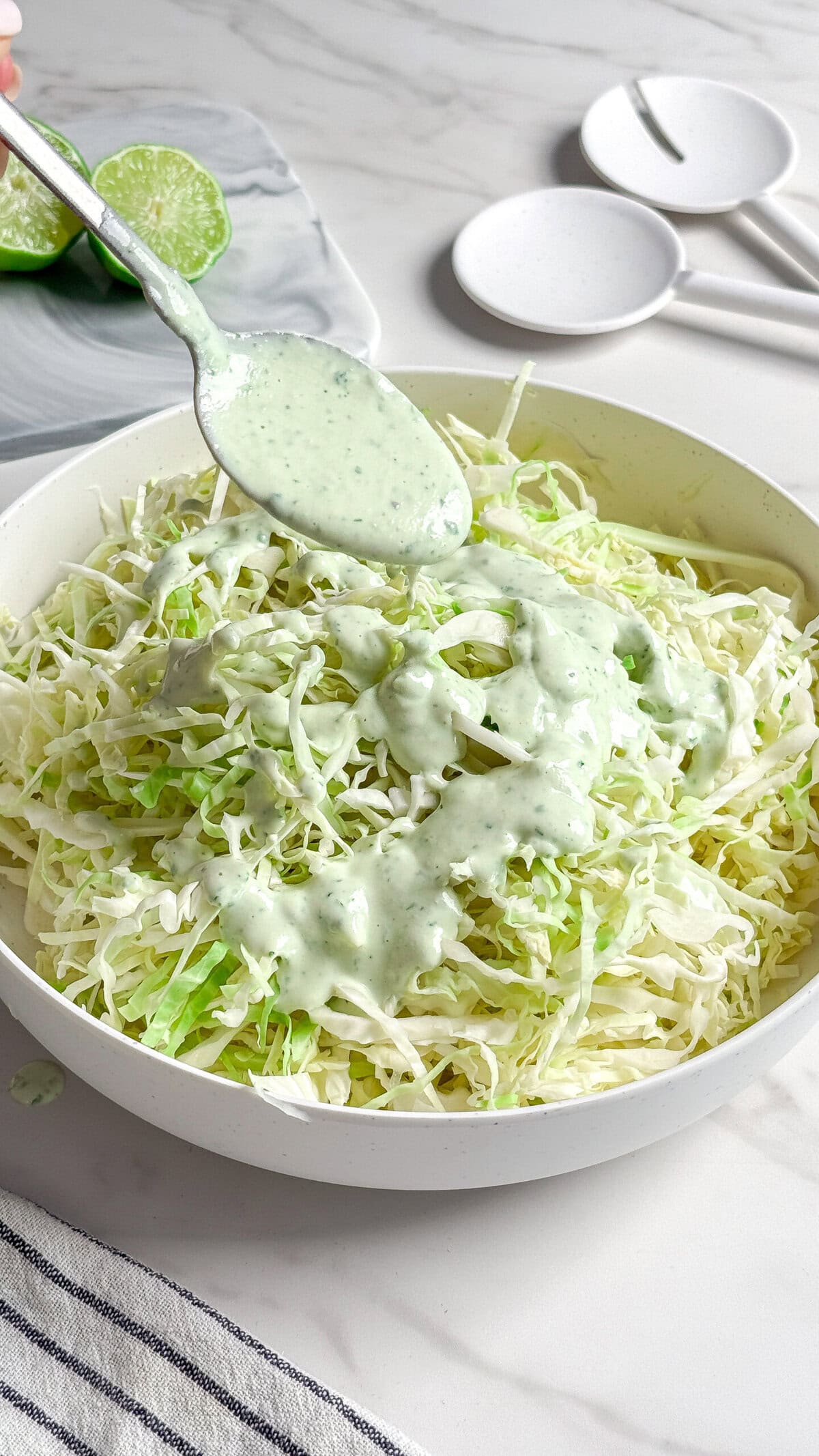 A bowl of shredded cabbage is being topped with a creamy green herb no mayo coleslaw dressing, spooned from above. A sliced lime and empty white condiment dishes are visible in the background on a marble surface.