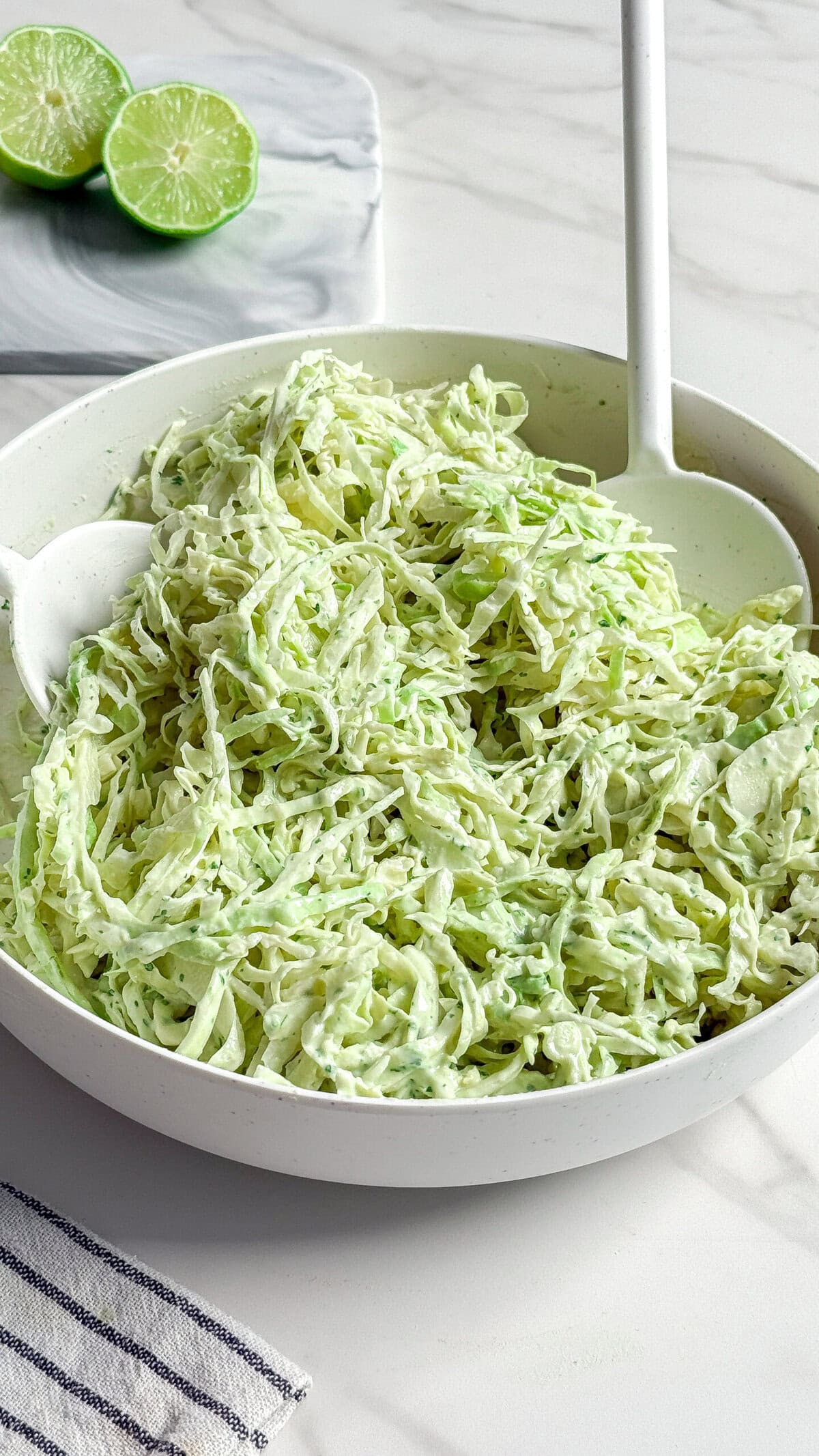 A white bowl filled with creamy no mayo coleslaw made from shredded cabbage, with a serving spoon inside. In the background, two halved limes rest on a marble surface. A striped cloth is partially visible in the corner.
