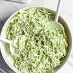 A white bowl filled with creamy no mayo coleslaw sits on a marble surface, with two white serving utensils in it. Nearby, there is a halved lime on a small tray and a striped cloth napkin.