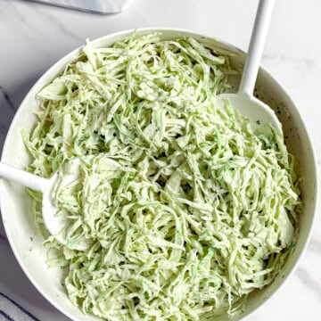 A white bowl filled with creamy no mayo coleslaw sits on a marble surface, with two white serving utensils in it. Nearby, there is a halved lime on a small tray and a striped cloth napkin.