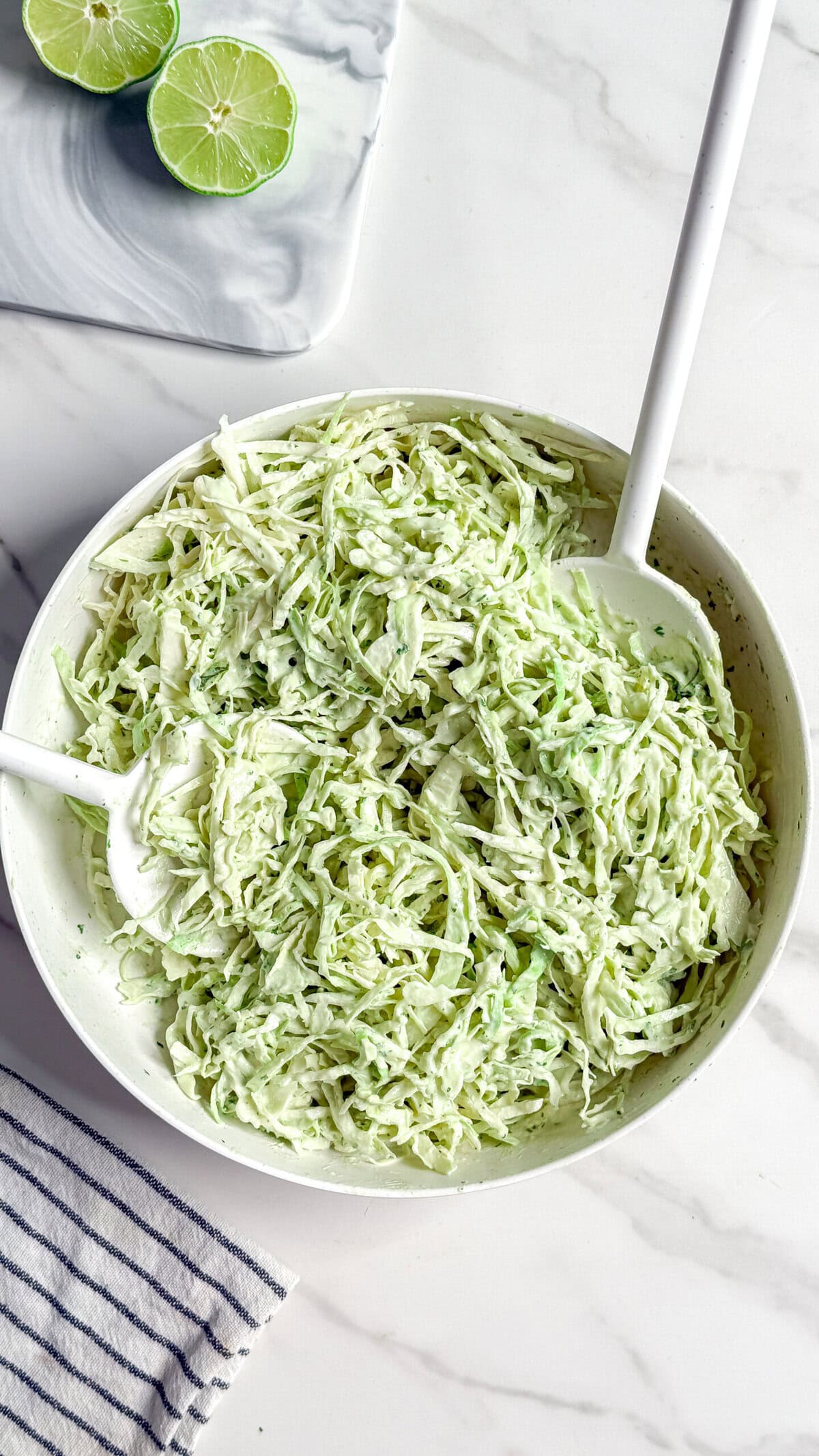 A white bowl filled with creamy no mayo coleslaw sits on a marble surface, with two white serving utensils in it. Nearby, there is a halved lime on a small tray and a striped cloth napkin.
