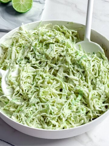 A white bowl filled with creamy no mayo coleslaw made of shredded cabbage and herbs sits on a marble surface, with a white serving spoon and sliced limes visible in the background.