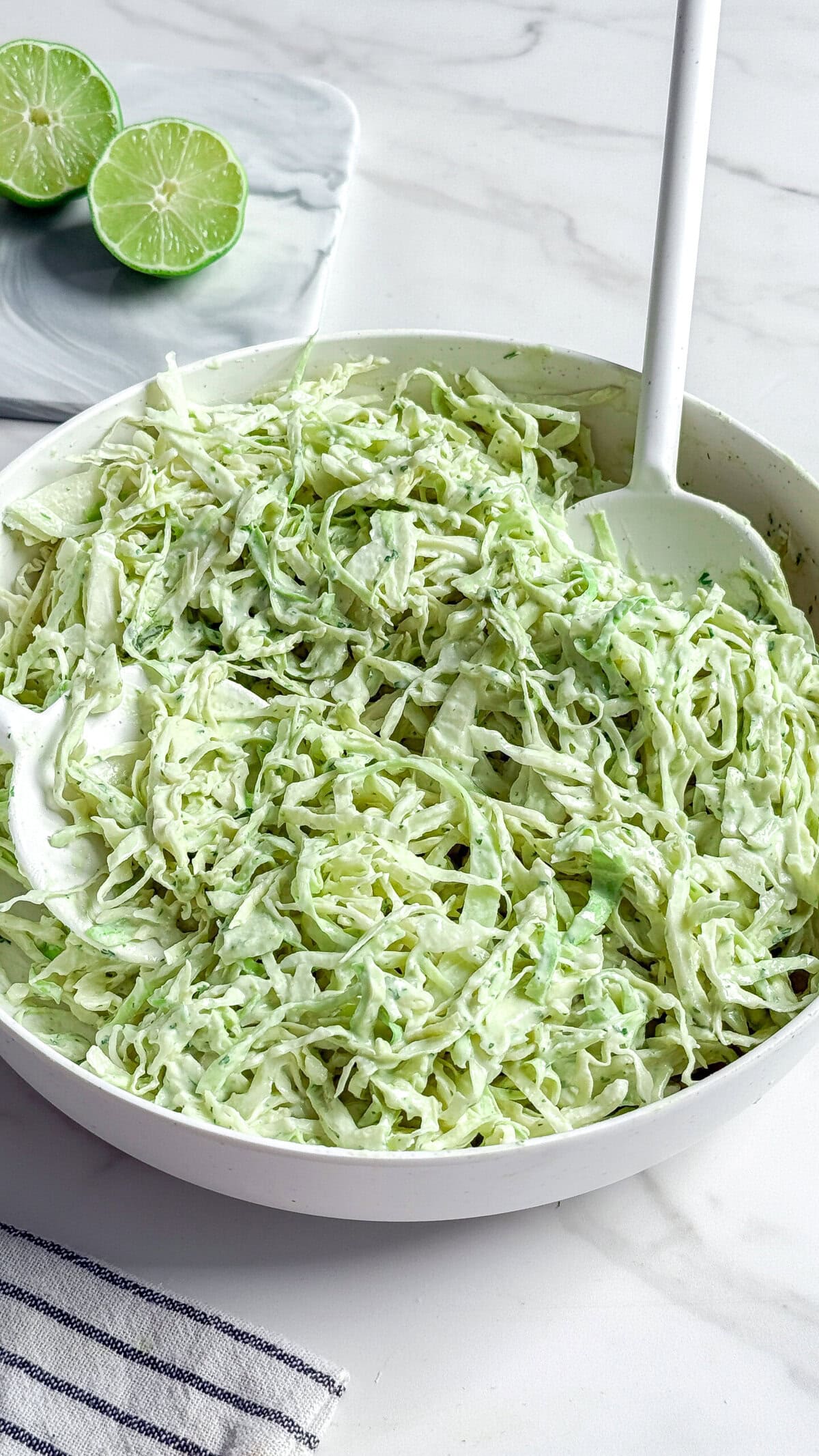 A white bowl filled with creamy no mayo coleslaw made of shredded cabbage and herbs sits on a marble surface, with a white serving spoon and sliced limes visible in the background.