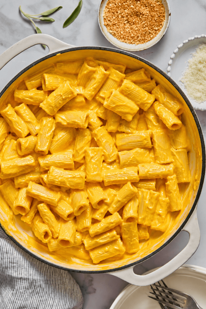 A pot filled with creamy rigatoni pasta coated in a rich, orange-yellow cheese sauce reminiscent of our signature Creamy Vegan Pumpkin Garlic Pasta. Bowls of breadcrumbs and grated cheese sit nearby on a marble surface.
