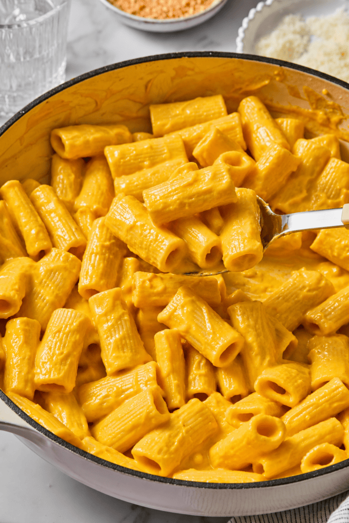 A pot filled with creamy rigatoni pasta coated in a rich orange cheese sauce, with a serving spoon lifting some pasta. In the background, there are bowls of grated cheese and crumbs. This dish is reminiscent of Creamy Vegan Pumpkin Garlic Pasta, blending comforting flavors and velvety texture in every bite.