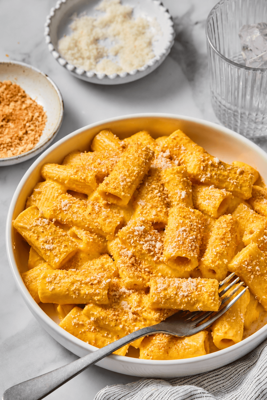 A bowl of rigatoni pasta covered in a creamy orange sauce, reminiscent of Creamy Vegan Pumpkin Garlic Pasta, topped with grated cheese and breadcrumbs, with a fork resting on the side. Dishes with extra cheese and breadcrumbs and a glass of ice water are nearby.