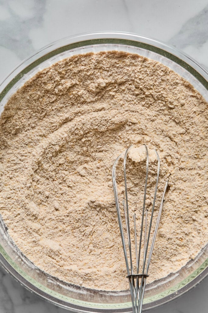 A metal whisk rests in a glass bowl filled with a light brown dry mixture, possibly flour or baking ingredients for a vegan gluten-free apple cake, on a marble surface.