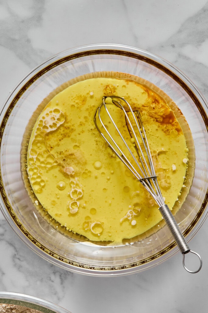 A glass bowl with liquid ingredients and a whisk, ready to be mixed for a vegan gluten-free apple cake, sits on a marble countertop. The mixture includes oil, eggs substitutes, and other wet ingredients.