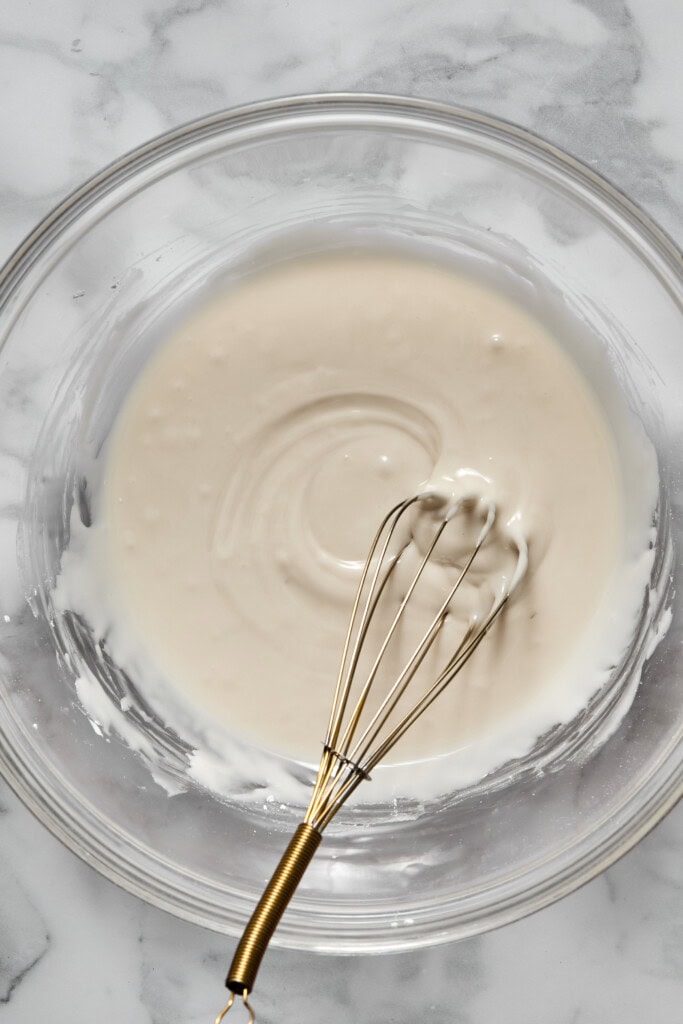A metal whisk rests in a glass bowl containing a smooth, light-colored batter for vegan gluten-free apple cake on a marble surface.
