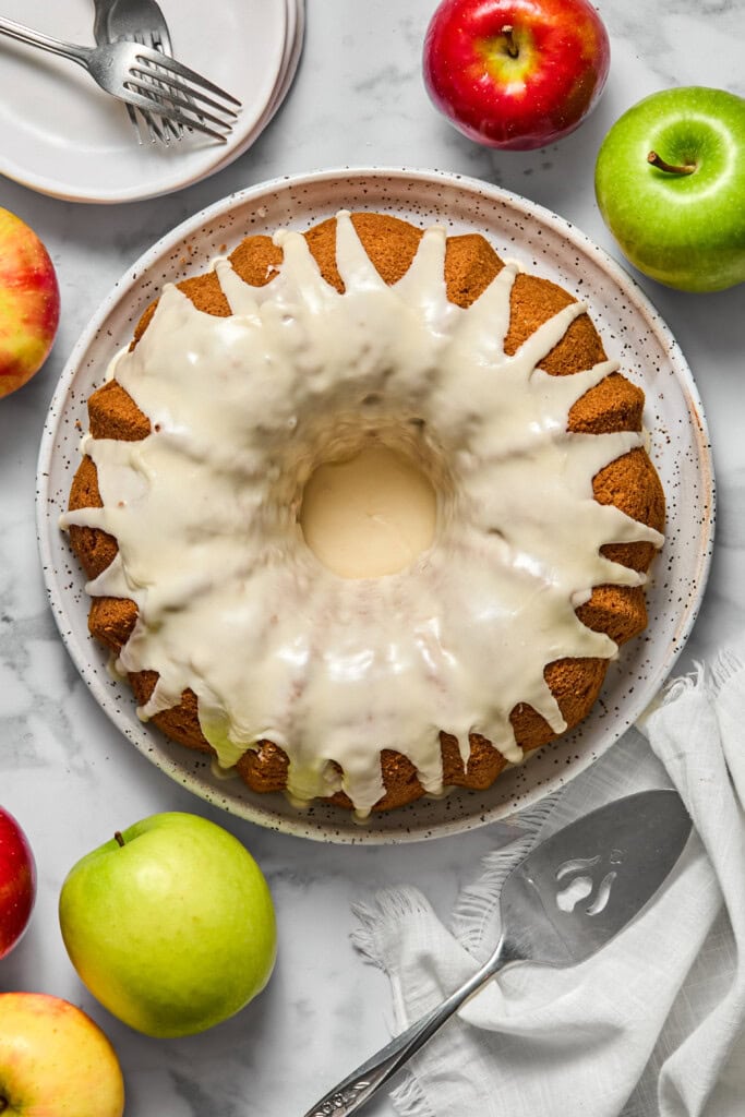 A glazed vegan gluten-free apple cake sits on a plate, surrounded by red and green apples, with plates, forks, and a pie server nearby on a marble surface.